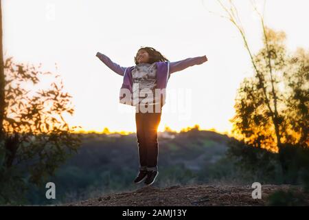 Ein junges Mädchen, das während eines Sonnenuntergangs mit Armen auf der Spitze eines Hügels sprang. Stockfoto