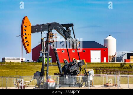 Eine Öl-Wells-Pumpenkäbe, oberirdische Hubkolbenpumpe, wird von einer großen Ranch in Alberta gesehen. Mit Getreidesilo, roter Scheune und Bauernhäusern im Hintergrund