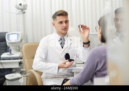 Portrait von gutaussehenden jungen Augenärzten, die Glaslinse halten, während sie mit weiblichen Patienten während der Beratung in der Klinik sprechen, Kopierraum Stockfoto
