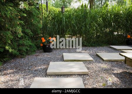 Eckige Zementpfadfliesen und schwarze Keramikfliesen mit orangefarbenen Begonienblüten und Thuja - Zedernbäume, Miscanthus sinensis - Ziergraspflanzen. Stockfoto