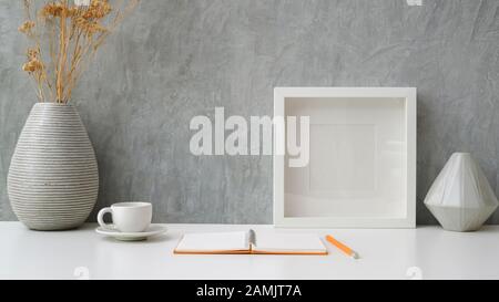 Nahaufnahme des modernen Arbeitsbereichs mit offenem Notizbuch, Kaffeetasse, Stehrahmen und Keramikvasen auf weißem Tisch mit grauem Loft-Wandhintergrund Stockfoto