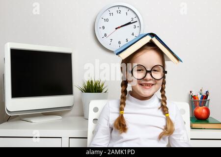 Eine fröhliche kleine Schülerin mit Brille sitzt in einem Raum neben einem Tisch mit einem Buch auf dem Kopf. Bildungs- und Schulkonzept. Stockfoto