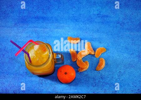 Ein Becher mit Tangerinsaft mit Eis und Strohhalmen befindet sich in einem Haufen Zitruspapieren auf blauem Grund. Nahaufnahme. Stockfoto