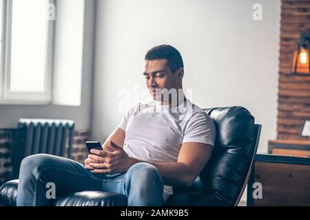 Junger Kerl in einem Stuhl, der auf ein Smartphone blickt. Stockfoto