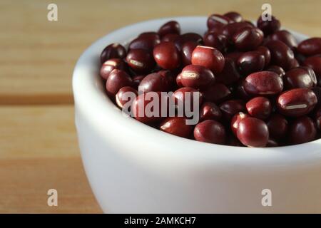 Eine schlichte, sonnige weiße Schüssel mit Adzuki-Bohnen, auch rote Mungbohne genannt, in Nahaufnahme auf einem einfachen Holztisch. Mit Copyspace. Stockfoto