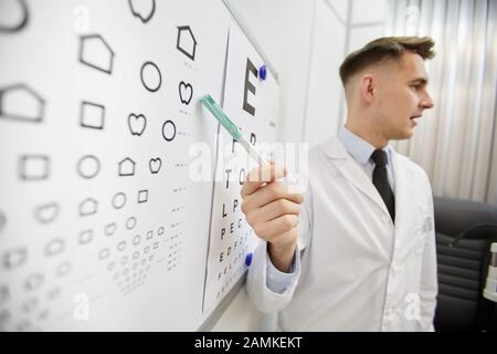 Seitenansicht des jungen Augenarztes, das auf die Vision-Chart zeigt, während das Augenlicht des nicht erkennbaren Patienten überprüft wird, Kopierbereich Stockfoto