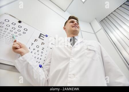 Das niedrige Porträt des lächelnden jungen Augenarztes, der auf das Vision-Diagramm zeigt, während er das Sehvermögen des nicht erkennbaren Patienten überprüft, den Kopierbereich Stockfoto