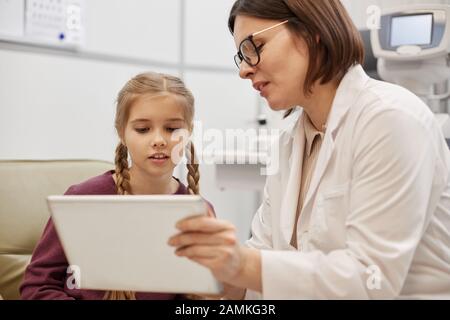 Portrait junger Augenärztin mit digitaler Tablette, während sie mit kleinen Mädchen während der Beratung in der modernen Klinik spricht Stockfoto