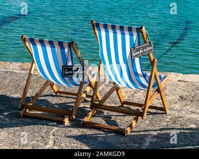 Zwei leere aber reserviert blau-weiß gestreifte Liegestühle mit' für Kaffee-/Zeichen" im Sommer, am Hafen von St. Ives, Cornwall, England, UK Gegangen Stockfoto