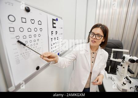 Hochwinkel-Porträt der weiblichen Augenärztin, die auf die Vision-Chart zeigt, während die Sehkraft des nicht erkennbaren Patienten überprüft wird, Kopierraum Stockfoto