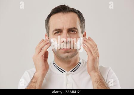 Kopf und Schultern Porträt des Mannes mittleren Alters, der Rasiercreme aufsetzt und während der morgendlichen Routine Kamera vor weißem Hintergrund betrachtet