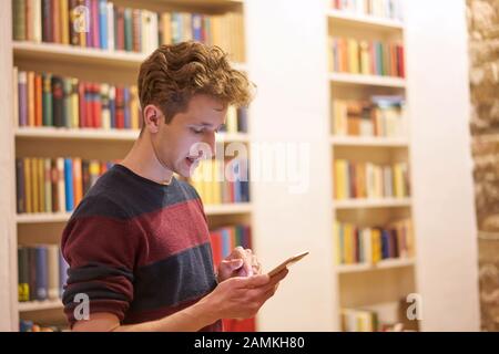 Junge konzentriert Student in Bibliothek nutzt sein Handy vor Bücherregal Stockfoto