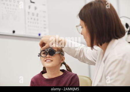 Portrait der weiblichen Optometristin, die einen Probelahmen aufstellt, während sie das Sehvermögen des kleinen Mädchens in der modernen Klinik für Augenheilkunde überprüft, kopiere Raum Stockfoto