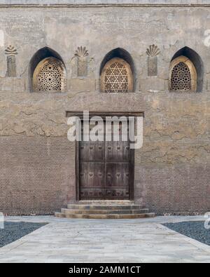 Verwitterte Holztür, perforiertes, mit Blumenmustern verziertes, gewölbtes Stuck-Fenster und drei Stufen auf Steinziegelwand, Außenfassade der Ibn-Tulun-Moschee, Kairo, Ägypten Stockfoto