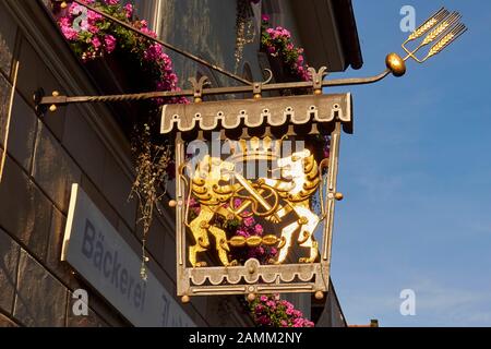 Goldenes Zunfzeichen einer Bäckerei in Bad Tölz, Krone, Löwen mit gekreuzten Schwertern und Brezel sowie drei Rollen, Balkone mit Blumenkästen, 12.10...2014, [automatisierte Übersetzung] Stockfoto