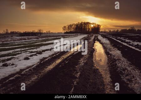 Schlammige, zugefrorene Landstraße durch Felder, Wolke und Sonnenuntergang Stockfoto