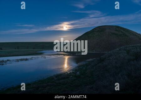 Ein Vollmond erhebt sich über dem riesigen neolithischen Kreidehügel von Silbury Hill, Avebury, Wiltshire, Großbritannien. Er ist 40 m hoch und wurde um 2300 v. Chr. erbaut Stockfoto