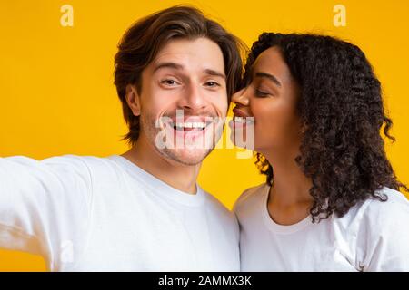 Fröhliche interracial Liebhaber, die selfie zusammen nehmen, schwarzes Mädchen, das ihren Freund küsst Stockfoto