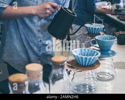 Barista in blauem Hemd, das heißes Wasser aus schwarzem Wasserkocher in den Kaffeeplatz mit Filter an der weißen Theke im Café gießt. Stockfoto