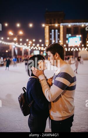 Das junge Paar skiert auf der Eisbahn und umarmt den Hintergrund der abendlichen Stadtbeleuchtung. Stockfoto