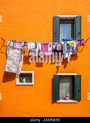 Trocknende Kleidung am Seil an der orangefarbenen Wand mit zwei Fenstern mit Fensterläden im Haus in Burano, Venedig. Stockfoto