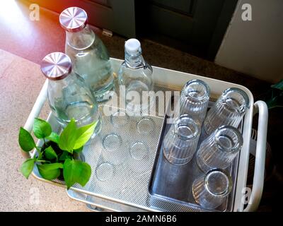 Kostenloser Trinkwasser-Selbstbedienung im Café. Glashaufen in Tablett und Trinkwasser-Flaschen auf Trolleydekoration mit grüner Anlage, Draufsicht. Stockfoto