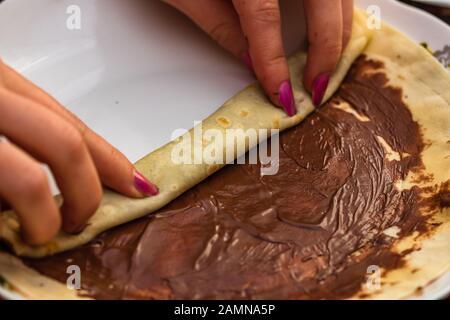 Hände bereiten Pfannkuchen mit Schokoladencreme vor und rollen, Nahaufnahme. Süßes hausgemachtes Dessert. Stockfoto