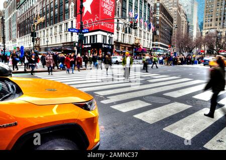 Selektive Fokussierung auf ein gelbes Taxi. Lange Exposition von Menschen, die die Straße überqueren. Bewegung, verschwommene Menschen. 6th Avenue. Macy's Store Stockfoto