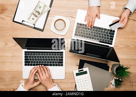 Draufsicht über Geschäftspartner, die Laptops mit Kreditkarte nach Dollarscheinen auf Holztisch verwenden, 3/4-Ansicht Stockfoto