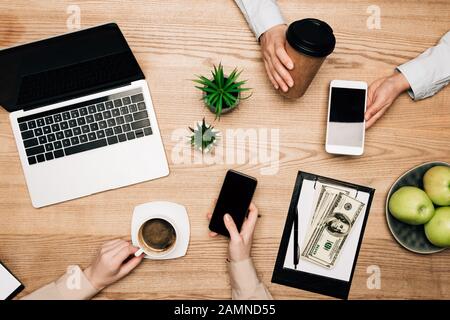 Draufsicht über Geschäftspartner, die Kaffee trinken und Smartphones mit Laptop und Geld auf dem Tisch nutzen Stockfoto