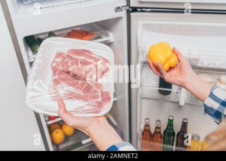 3/4-Ansicht der Frau, die gefrorenes Fleisch und Pfeffer in der Nähe eines offenen Kühlschranks hält, mit frischen Lebensmitteln in den Regalen Stockfoto