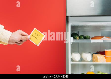 Beschnittener Blick auf den Mann, der klebrige Note mit guten Morgenschriften in der Nähe eines offenen Kühlschranks hält, mit frischen Speisen in den Regalen, die auf Rot isoliert sind Stockfoto