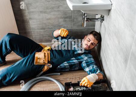 Bärtiger Handwerker, der auf dem Boden in der Nähe der Werkzeugkiste im Badezimmer liegt Stockfoto