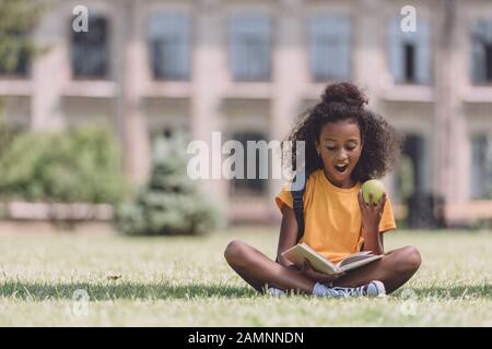 Überraschte afroamerikanische Schulmädchen, die Buch lesen, während sie auf Rasen sitzen und Apple halten Stockfoto