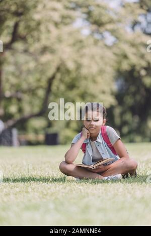 Süße, gelangweilte Schulmädchen, die Buch halten, während sie auf dem Rasen im Park sitzen Stockfoto