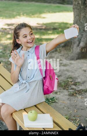 Fröhliches Schulmädchen winkt beim Sitzen auf der Bank die Hand und nimmt selfie mit Smartphone Stockfoto