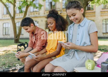 Drei niedliche multikulturelle Schüler, die Smartphones benutzen, während sie auf der Bank im Schulhof sitzen Stockfoto