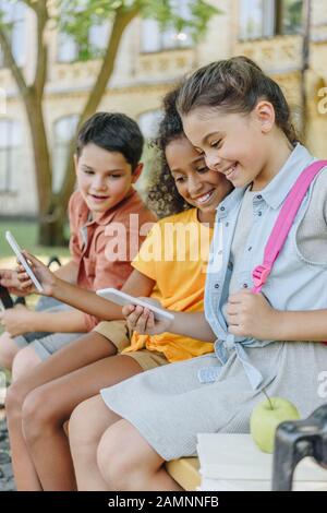 Drei fröhliche multiethnische Schüler, die Smartphones benutzen, während sie auf der Bank im Schulhof sitzen Stockfoto