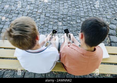 Overhead-Ansicht von zwei Schuljungen, die auf der Bank sitzen und Smartphones verwenden Stockfoto