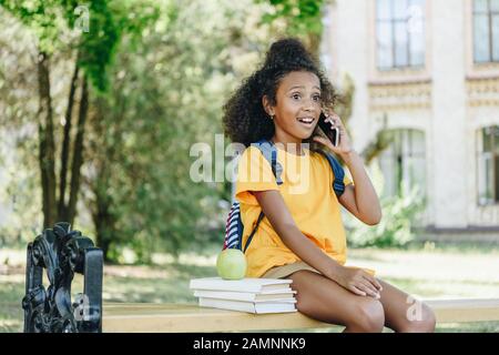 Überraschte das afroamerikanische Mädchen, das auf dem Smartphone spricht, während es auf der Bank in der Nähe von Büchern und apple sitzt Stockfoto