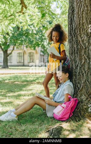Wunderbares afroamerikanisches Schulmädchen, das in der Nähe eines Freundes steht, der unter Bäumen auf dem Rasen sitzt und Buch liest Stockfoto