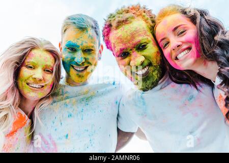 Die untere Ansicht von fröhlichen multikulturellen Freunden mit bunten Holi-Farben auf den Gesichtern Stockfoto