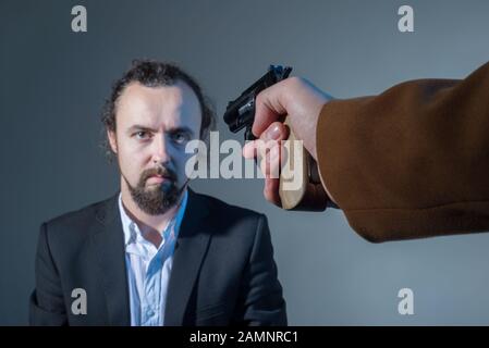 Nahaufnahme eines Mannes, dessen Gesicht mit einer Waffe eines anderen Mannes eine Hand anstrebt. Auf grauem Hintergrund. Tötungskonzept. Dramatisches Bild. Business di Stockfoto