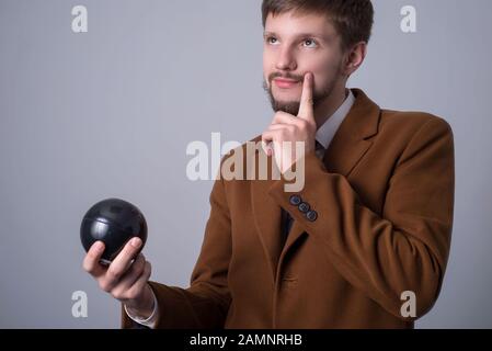 Porträt eines bärtigen Mannes in einem Business-Anzug, hält einen Ball für Wünsche und bittet einen schwarzen Billardball, der seine Hand in der Nähe seines Kinn hält, Träume, Stockfoto