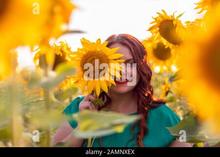 Porträt eines schönen asiatischen jungen Mädchens mit roten lockigen Haaren und einem grünen Kleid, das im Sommer an einem sonnigen Tag auf einem Sonnenblumenfeld posiert. Freiheit Stockfoto