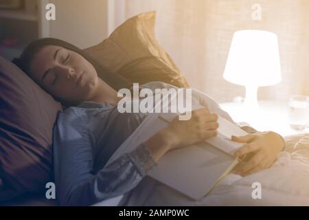 Junge Frau, die sich nachts in ihrem Bett erholt, schläft sie beim Lesen eines Buches ein Stockfoto