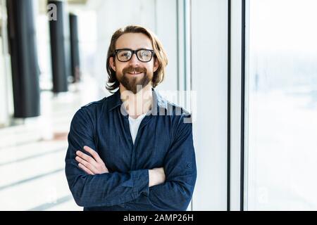 Selbstbewusster Kollege. Freudiger, gutaussehender Mitarbeiter in einer Brille, die im Büro in der Nähe des Tisches steht und seine Hände überkreuzt und lächelnd hält. Stockfoto