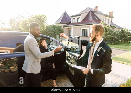 Sales Situation in ein Autohaus im Freien. Junge afrikanische Mann erhält den Schlüssel für das neue Auto von Kaukasischen sales manager. Aufgeregt, fröhliche Frau ist Stockfoto