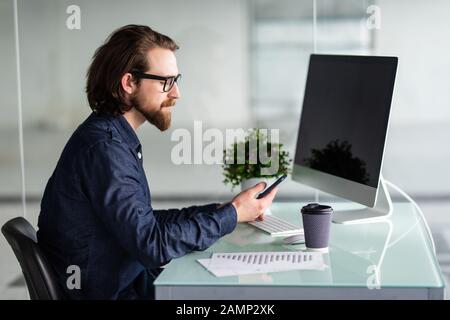 Der junge bärtige Geschäftsmann im Hemd sitzt im Büro am Tisch und verwendet Smartphone mit Karten, Diagrammen und Grafiken auf dem Bildschirm und Getränkekaffee. Stockfoto