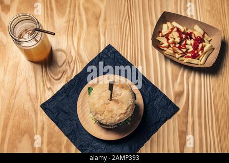 Draufsicht eines Gourmet-Hamburgers auf einem Holztisch mit pommes frites mit Ketchup in einem Pappkarton und einem gesunden Getränk, Fast-Food und Feinschmeckerlebensmittel Stockfoto